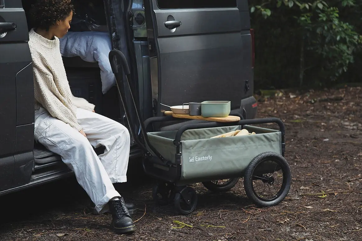 Persons sits on car in the woods next to green wagon set up for a picnic. 