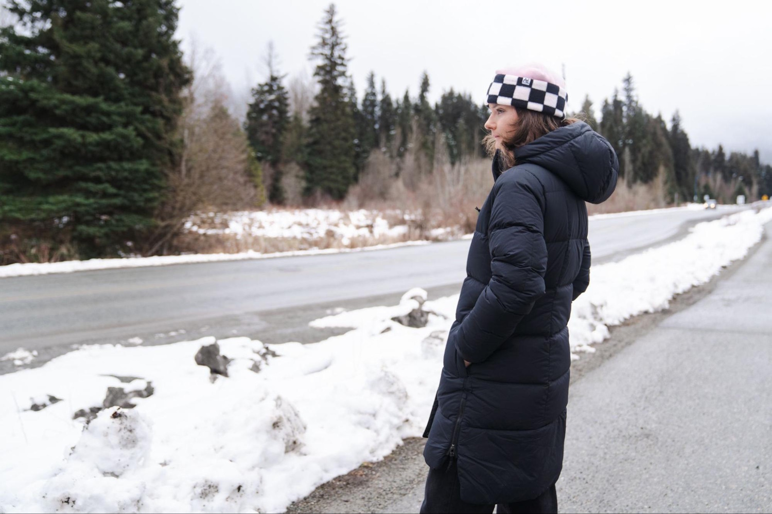 Woman wearing a long black parka stands by a snowy roadside