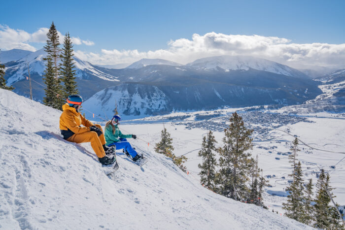 two snowboarders taking a break for the views at the top of a run overlooking Crested Butte town at Crested Butte Mountain Resort