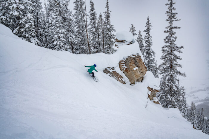 snowboarder takes steep turn beneath rock feature on a powder day