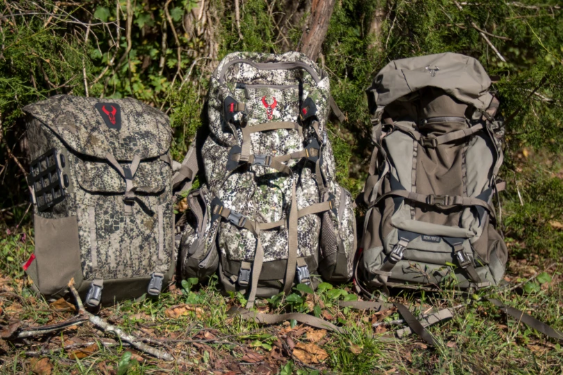 Three different sized hunting backpacks next to eachother in front of brush.