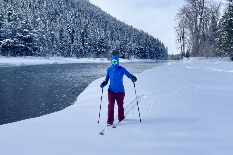 A person skis beside a calm river surrounded by snow-covered trees and mountains