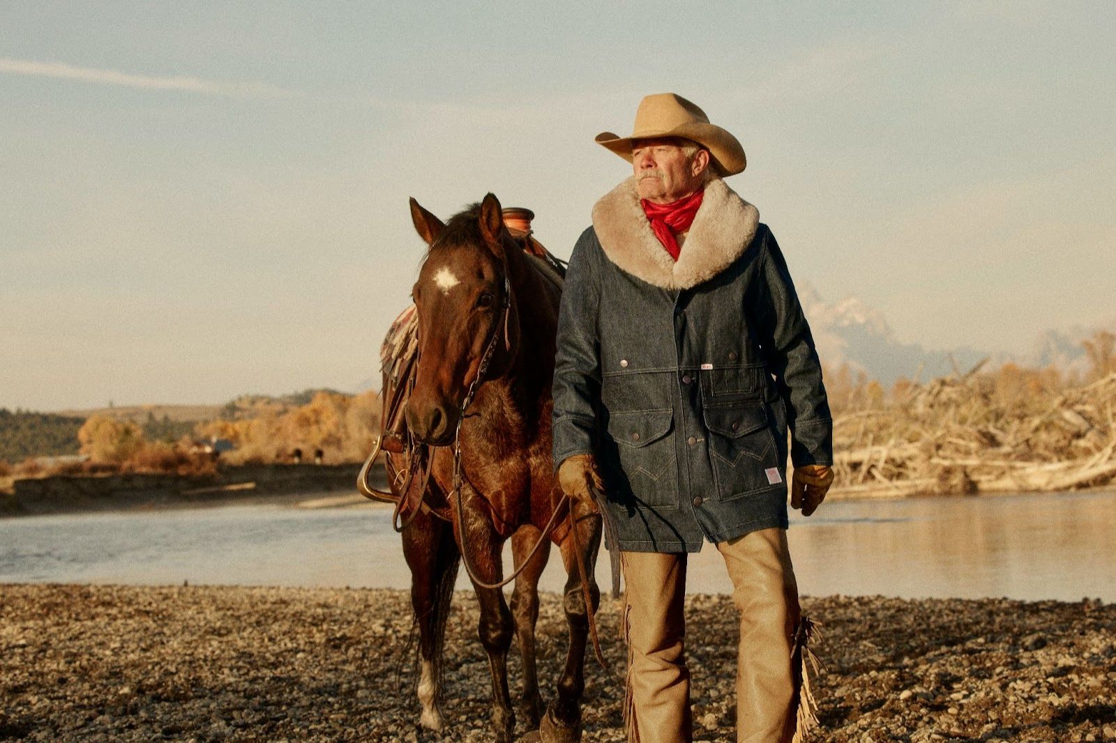 Cowboy in denim jacket walks next to lake with horse