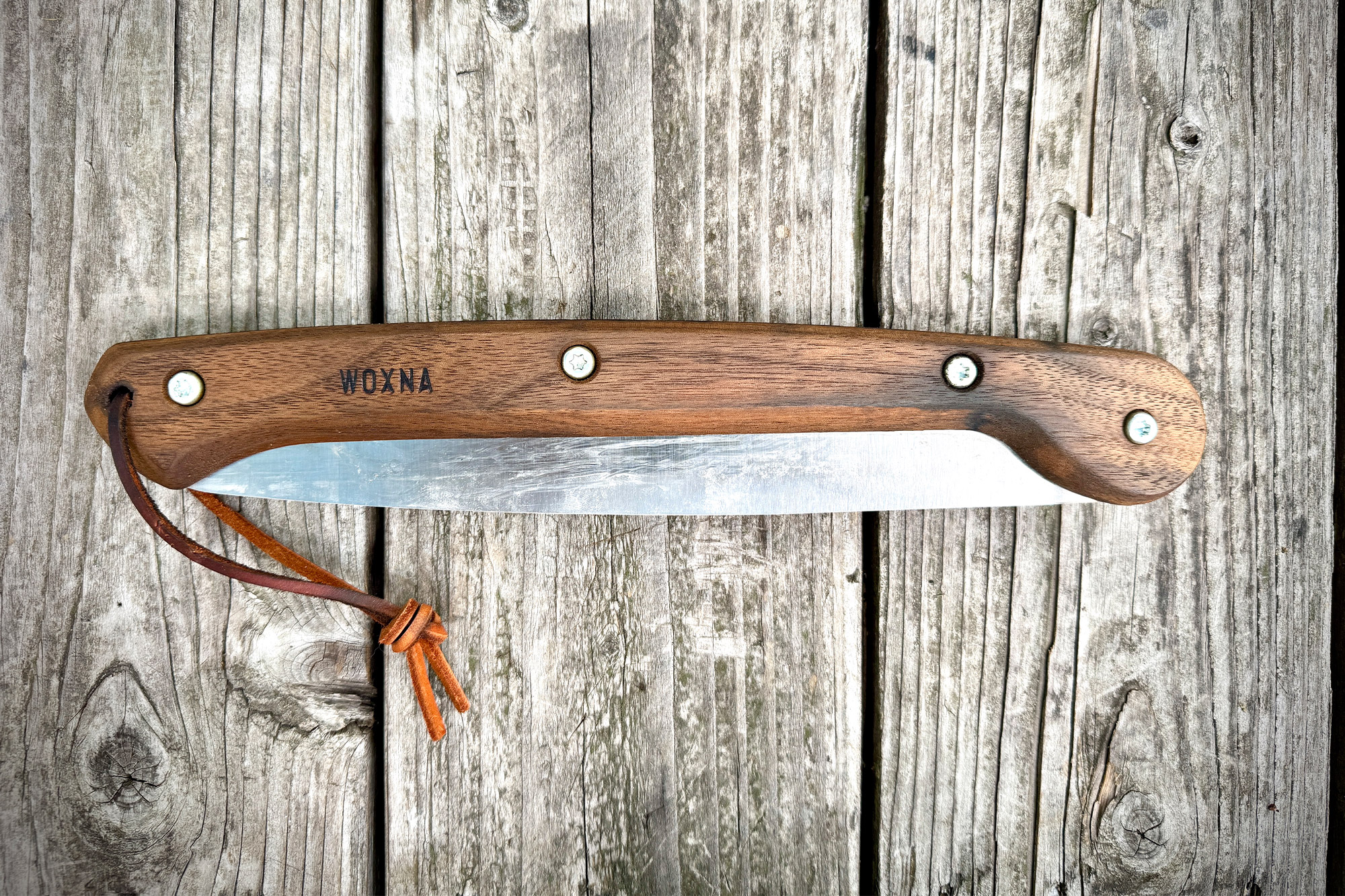 Woxna Ljusnan 10” Folding Saw shown folded, with a smooth wooden handle and leather strap resting on a weathered wood surface