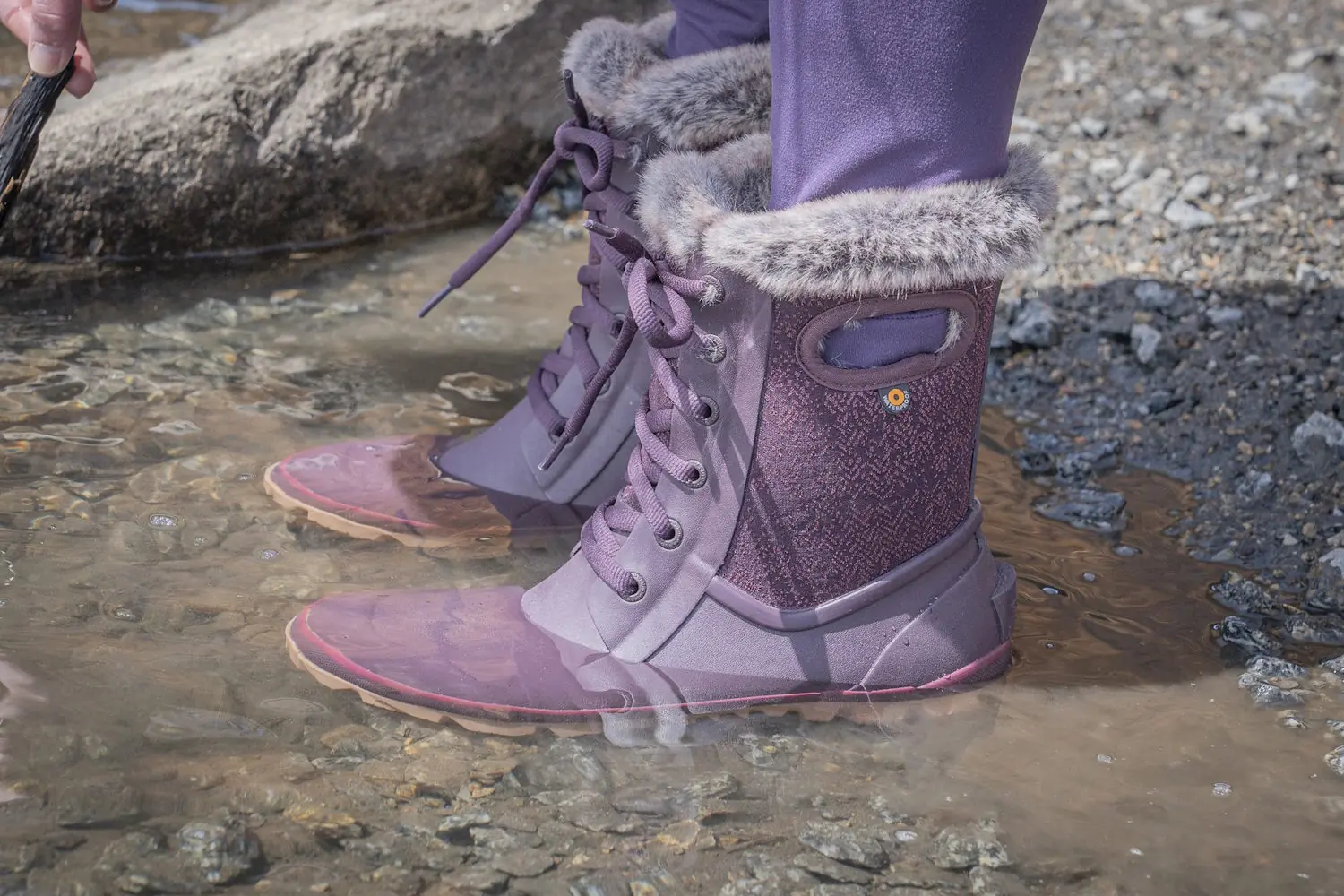 Close-up of purple Bogs Arcata Faded boots with faux fur lining, worn in a shallow rocky puddle