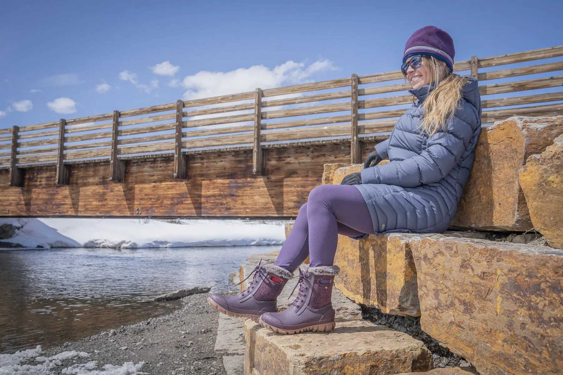 Woman wearing Bogs Arcata Faded winter boots sitting on stone steps beside a snowy river
