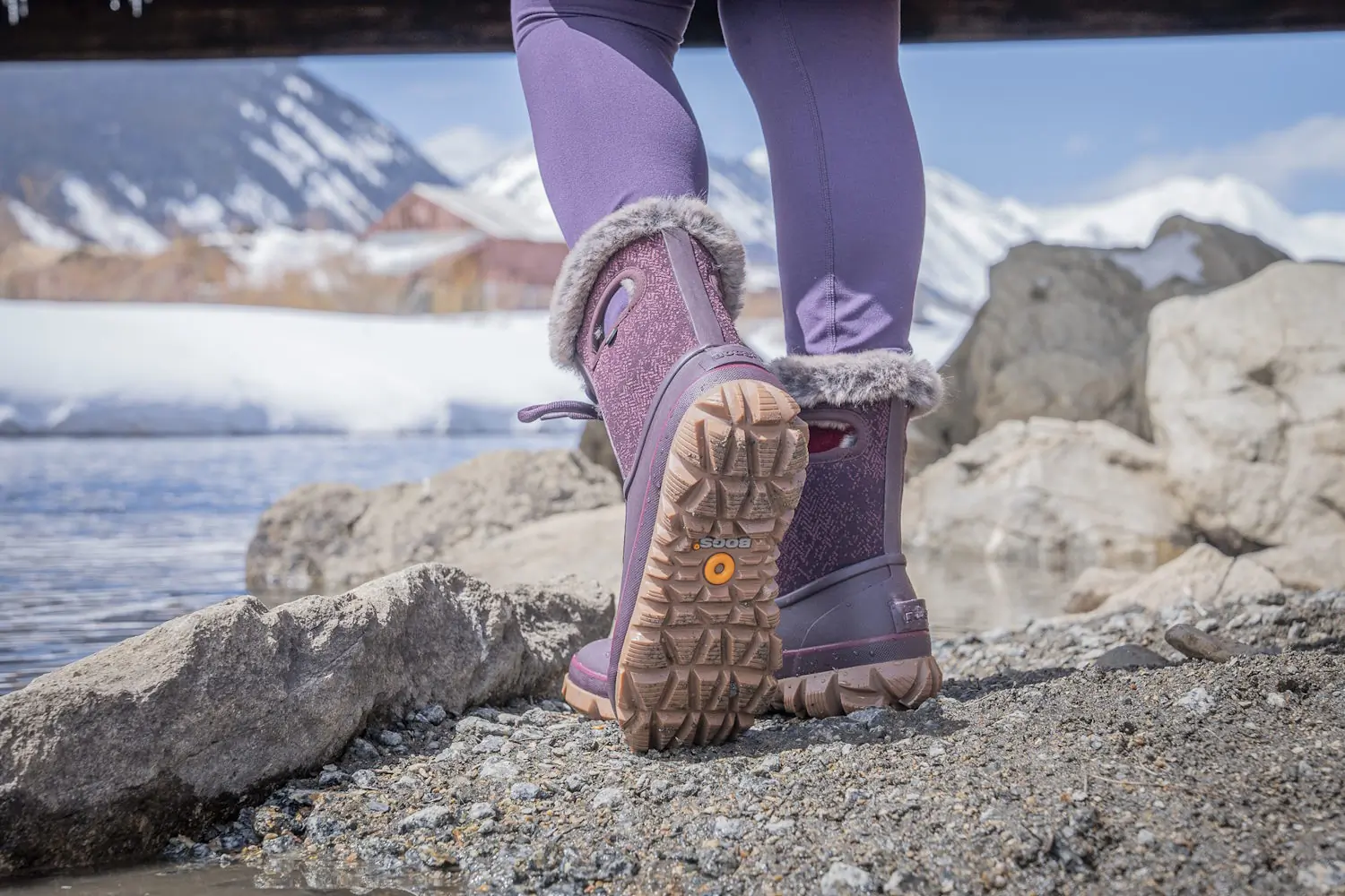 Close-up of a person wearing purple Bogs Arcata Faded winter boots standing on rocky, snowy terrain