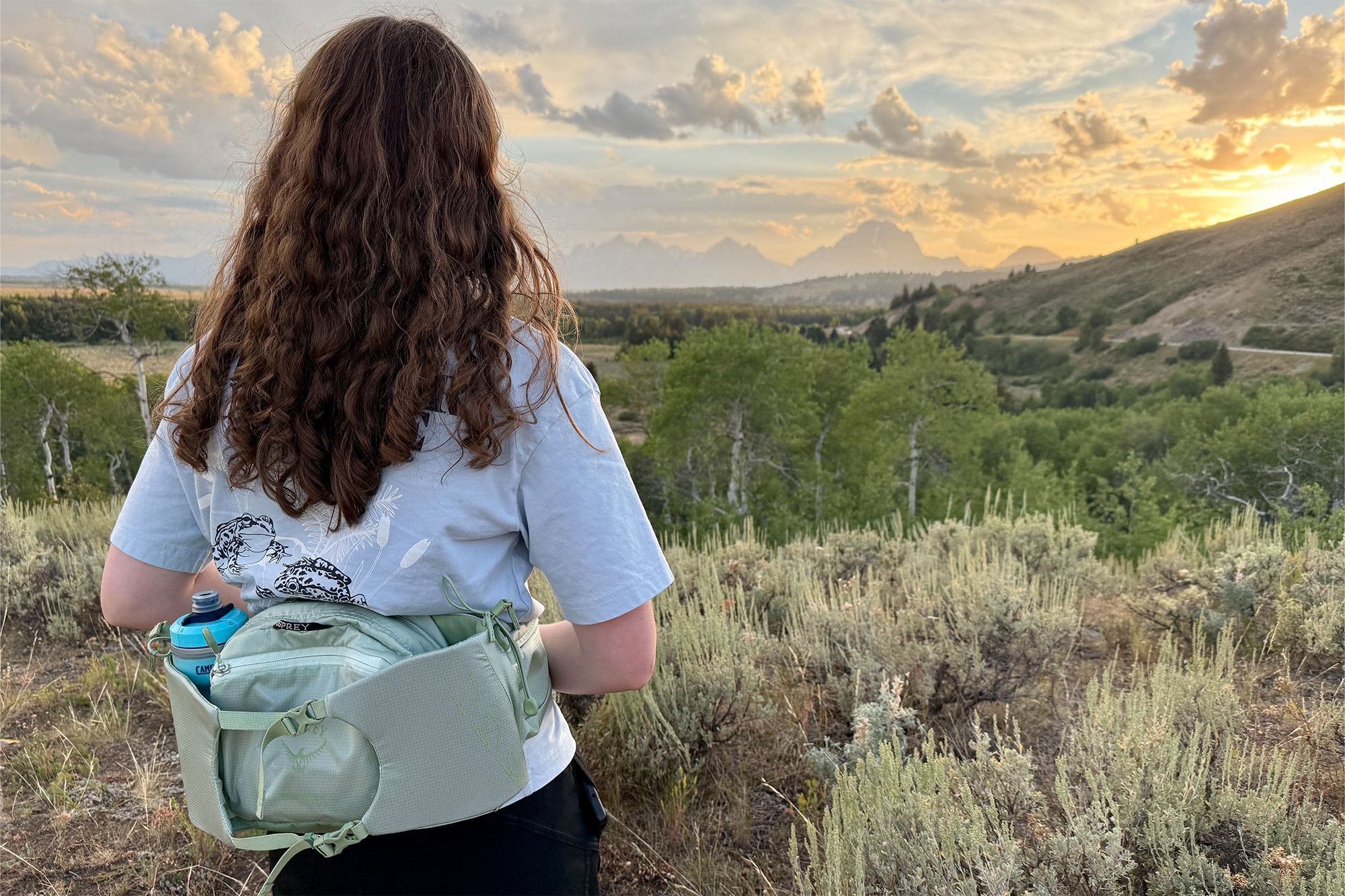 A person facing sunset views over mountain ridges, wearing the Osprey Tempest & Talon 6 Waistpack