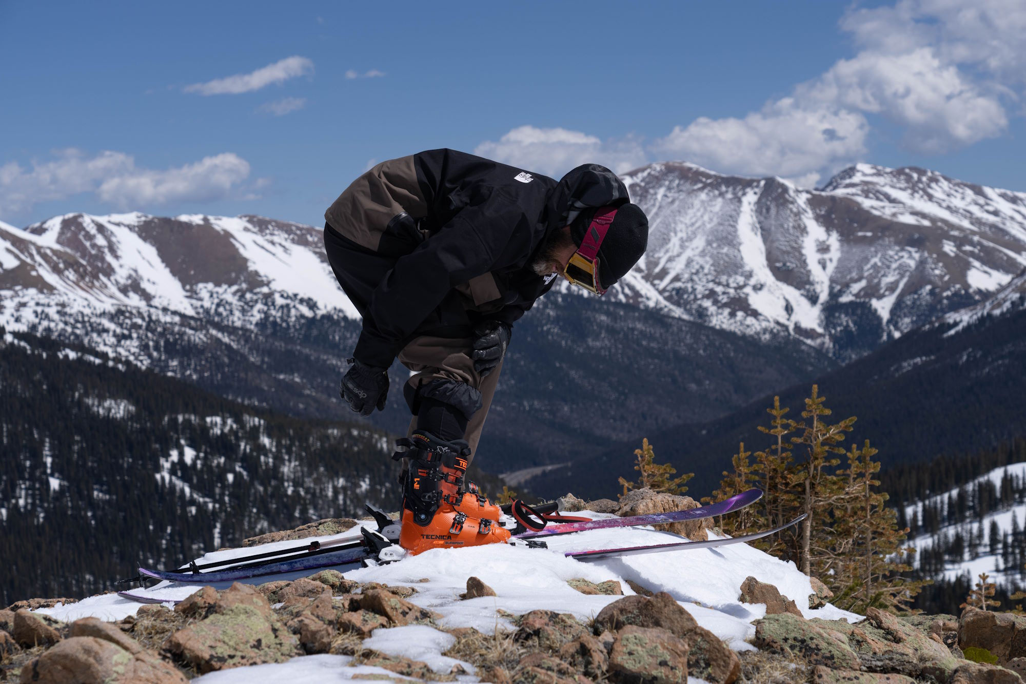Skier adjusting Tecnica Mach1 LV 130 boots atop a rocky, snow-covered ridge with alpine peaks in the background.