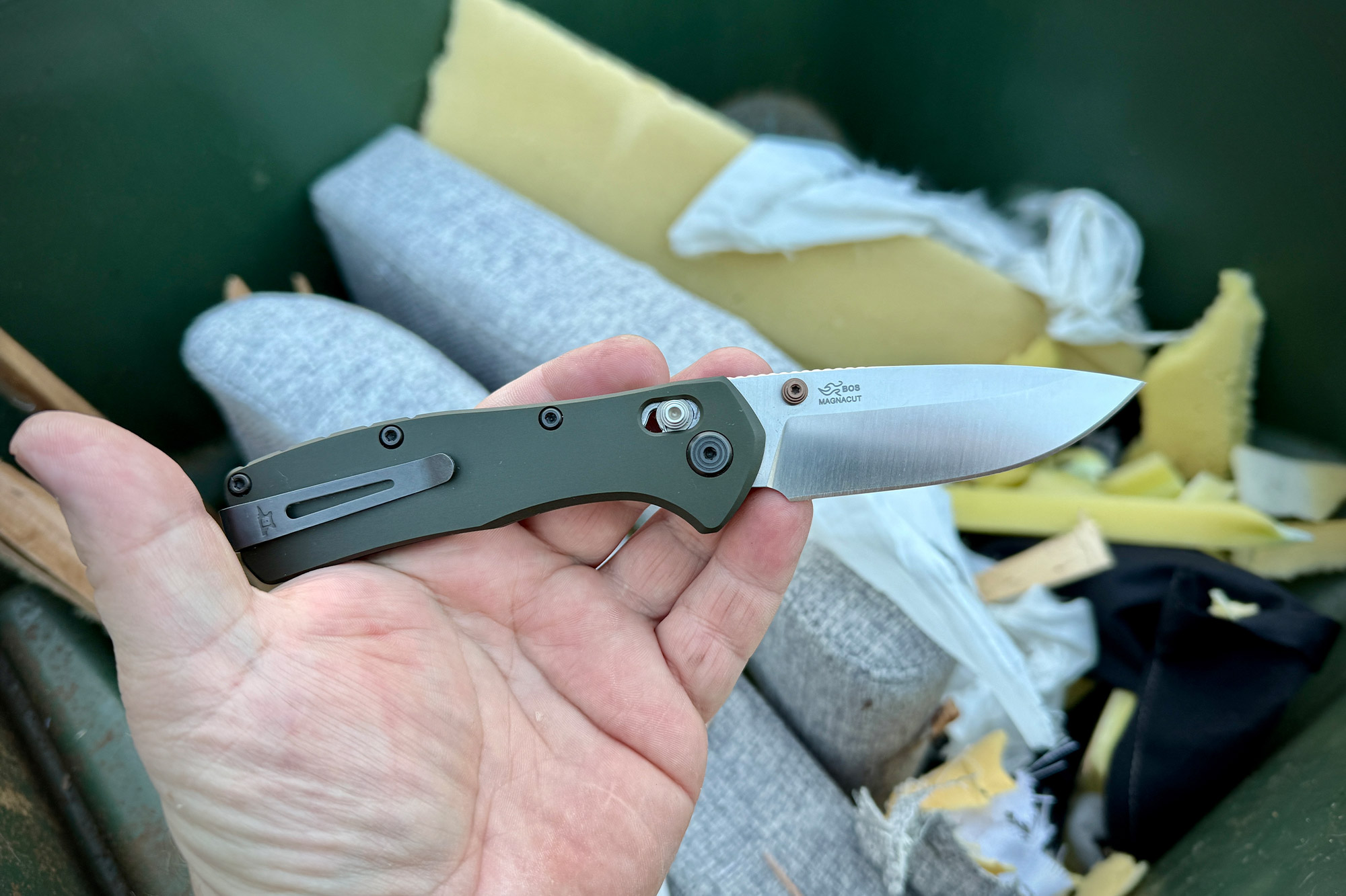 A Buck Range Elite knife held in a hand above torn upholstery inside a dumpster, blade open and clean