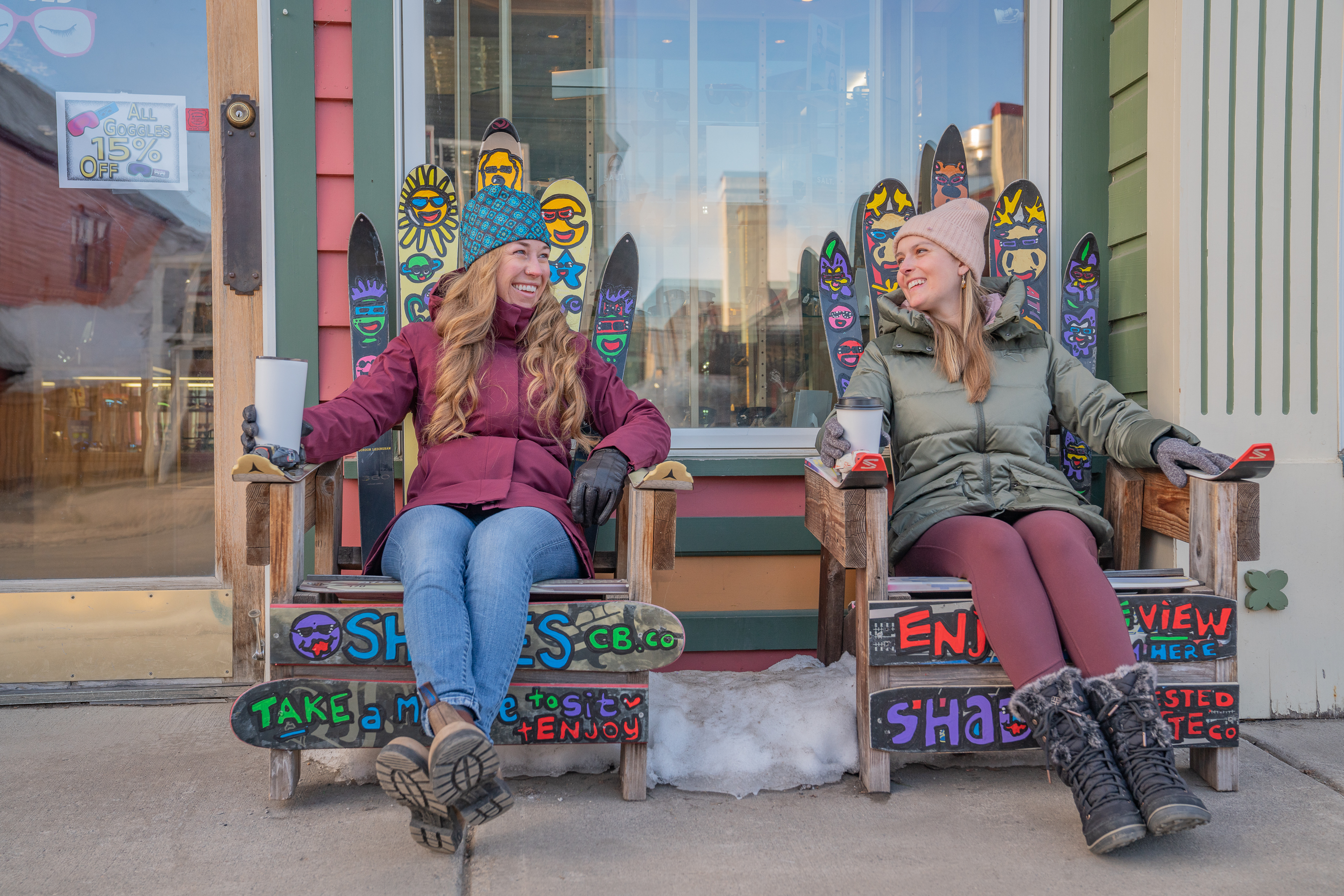 Two women wearing winter jackets and sitting in outdoor chairs at a storefront 