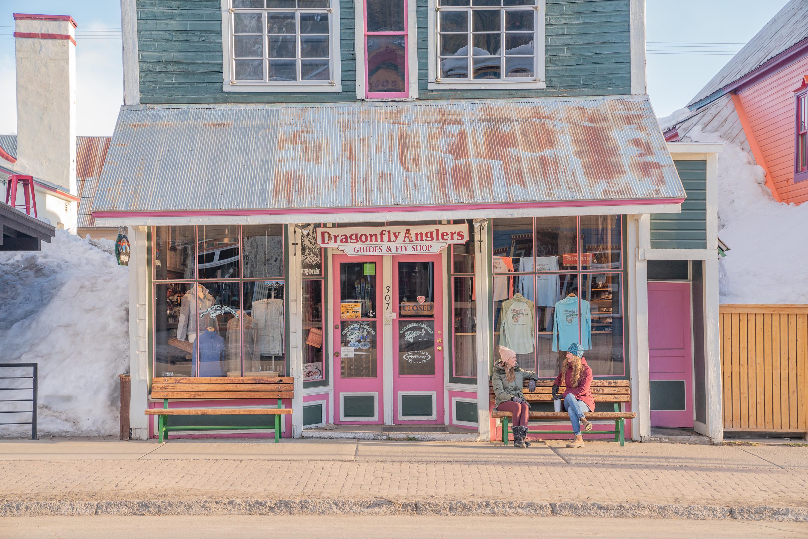 Two women in winter jackets sitting on a bench in front of a fly fishing shop 