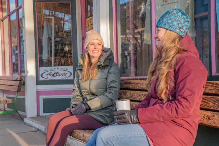 two women sitting on sidewalk bench testing winter jackets 