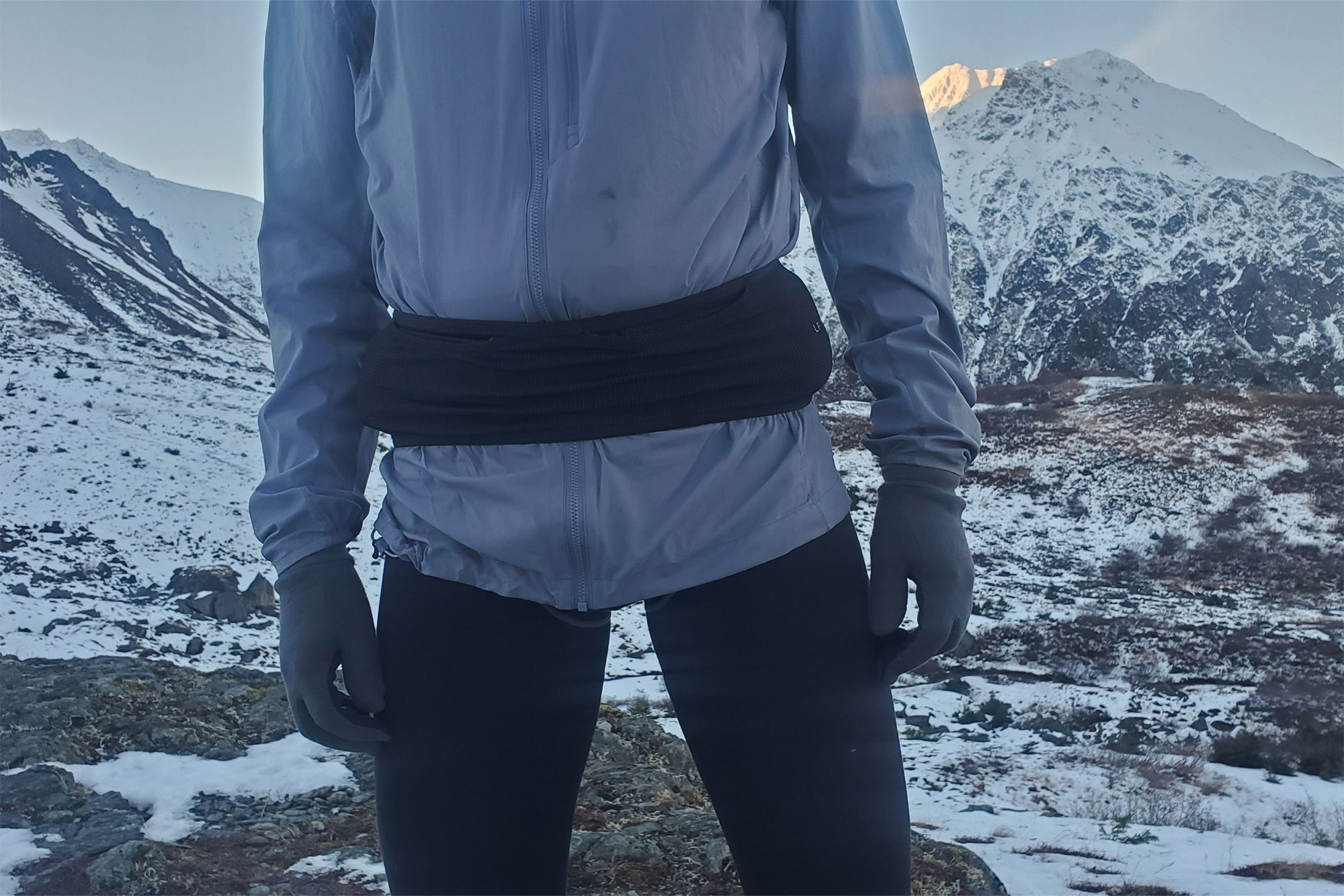 A person facing forward outdoors, wearing the running belt high on their waist over a gray jacket in a snowy mountain setting