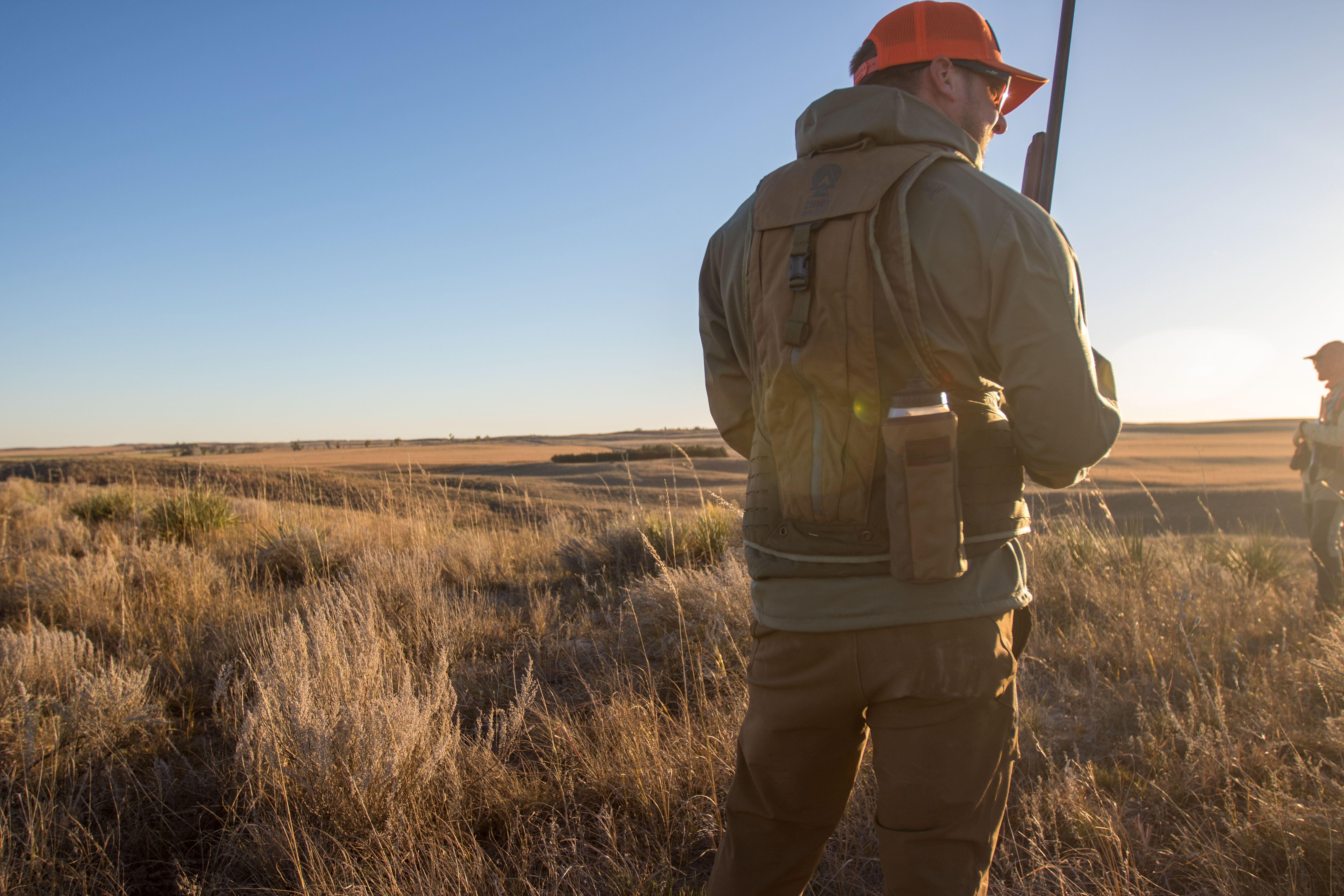 An upland hunter wearing a vest and holding a shotgun in the sunset.