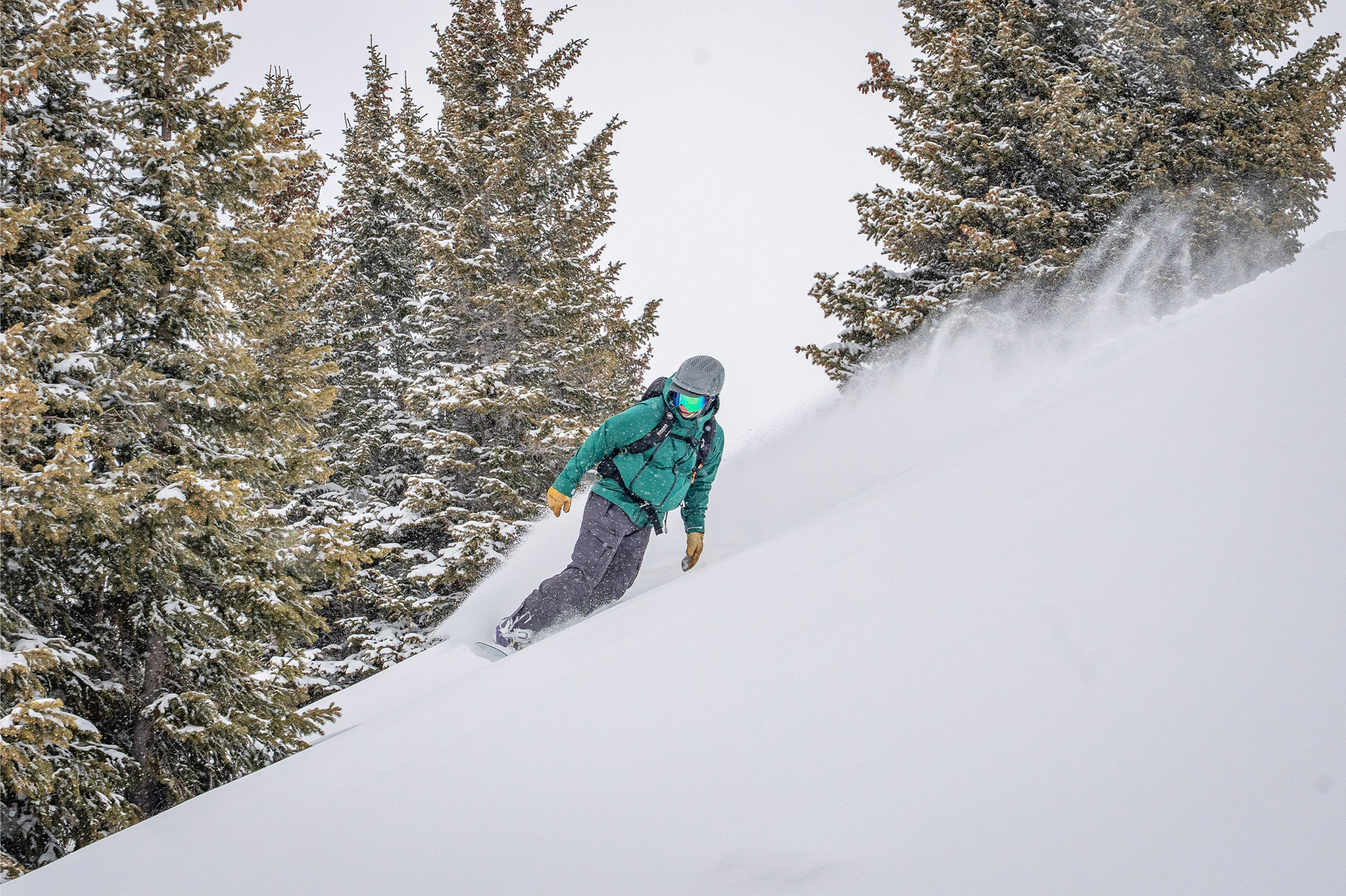 A rider carves through deep powder wearing the 686 snowboard jacket, spraying snow behind them