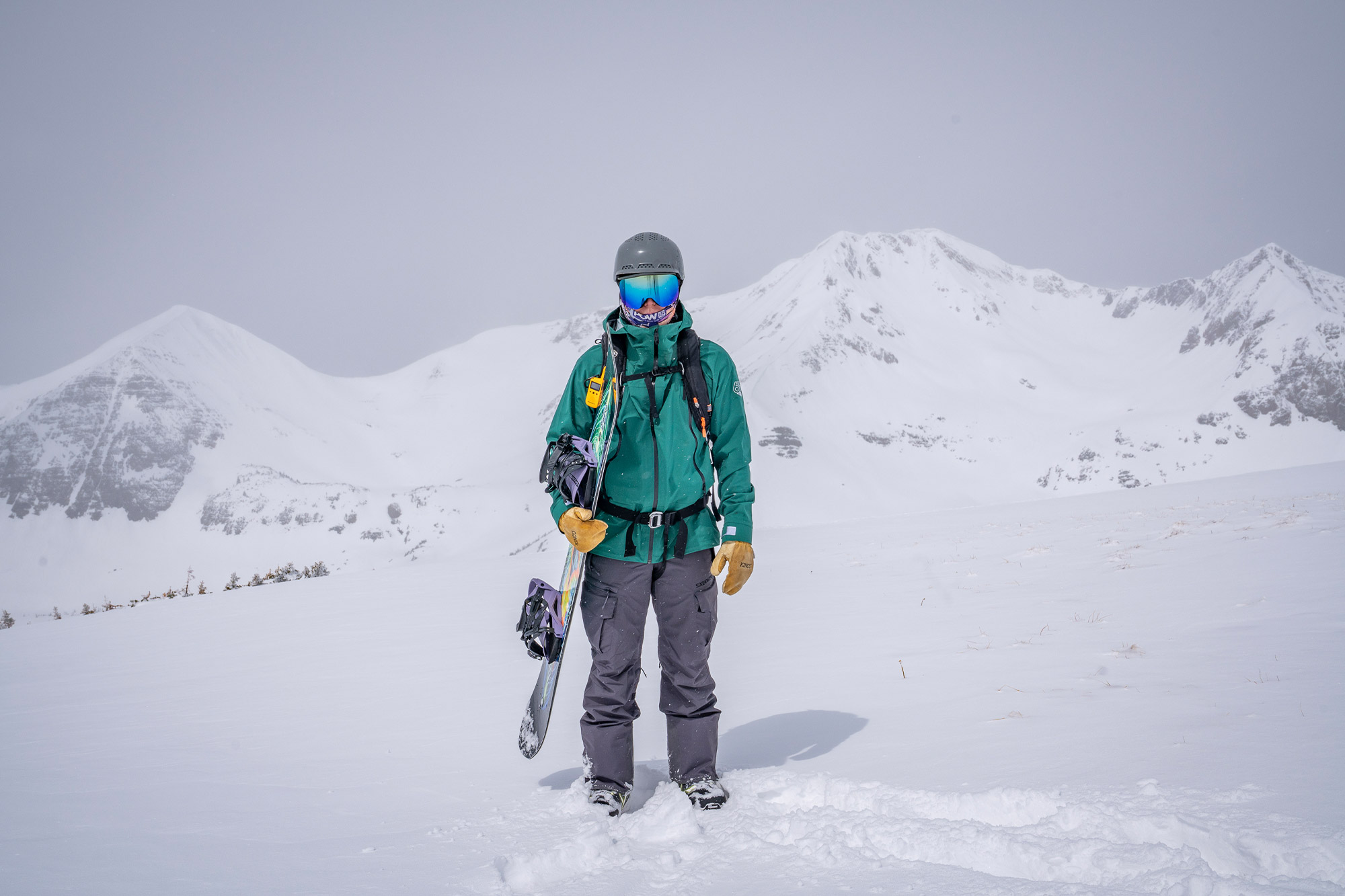 A snowboarder stands in an open snowy basin holding a board while wearing the 686 3-in-1 Rodeo Jacket