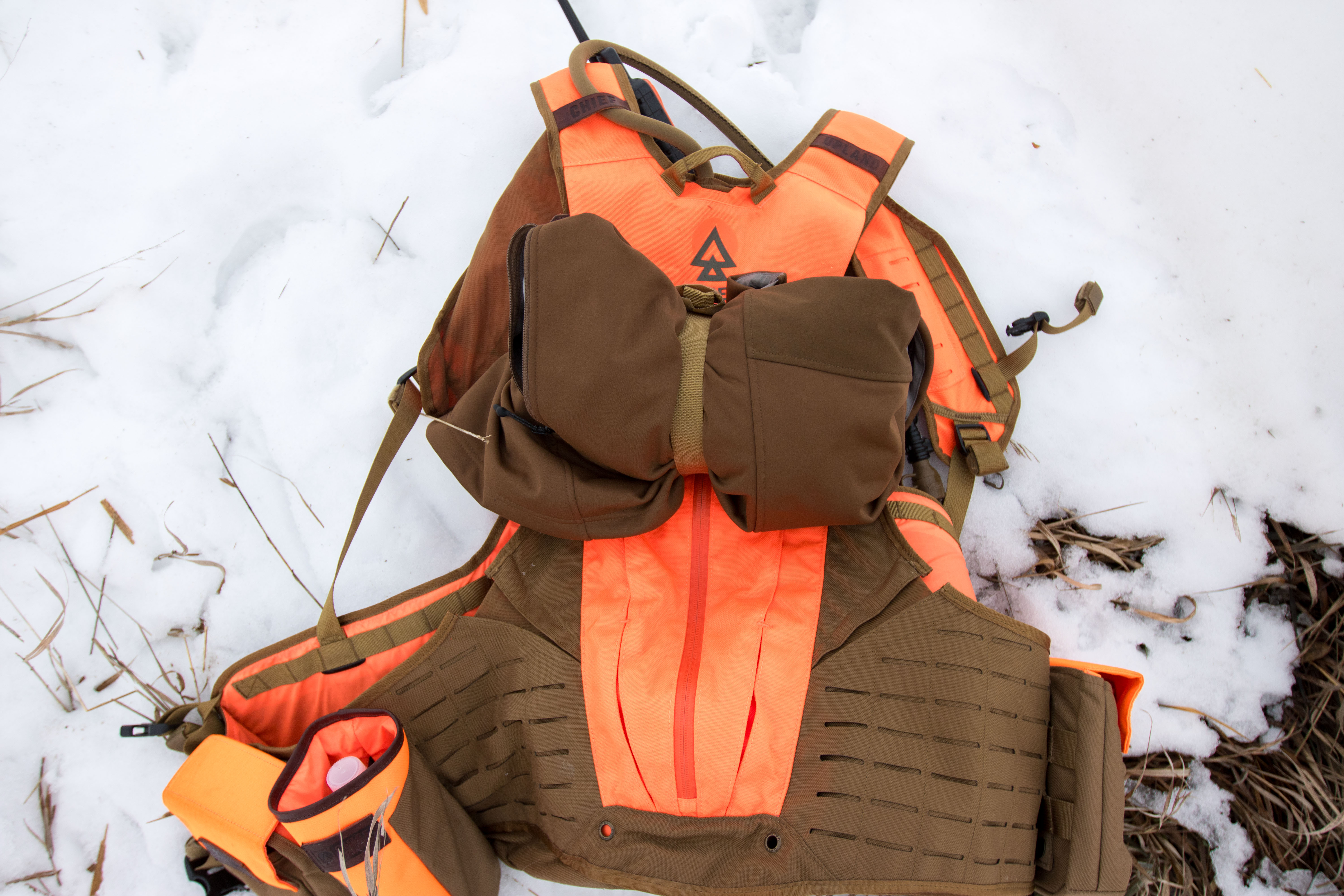 An upland bird hunting vest with a jacket strapped to the exterior, lying in the snow.
