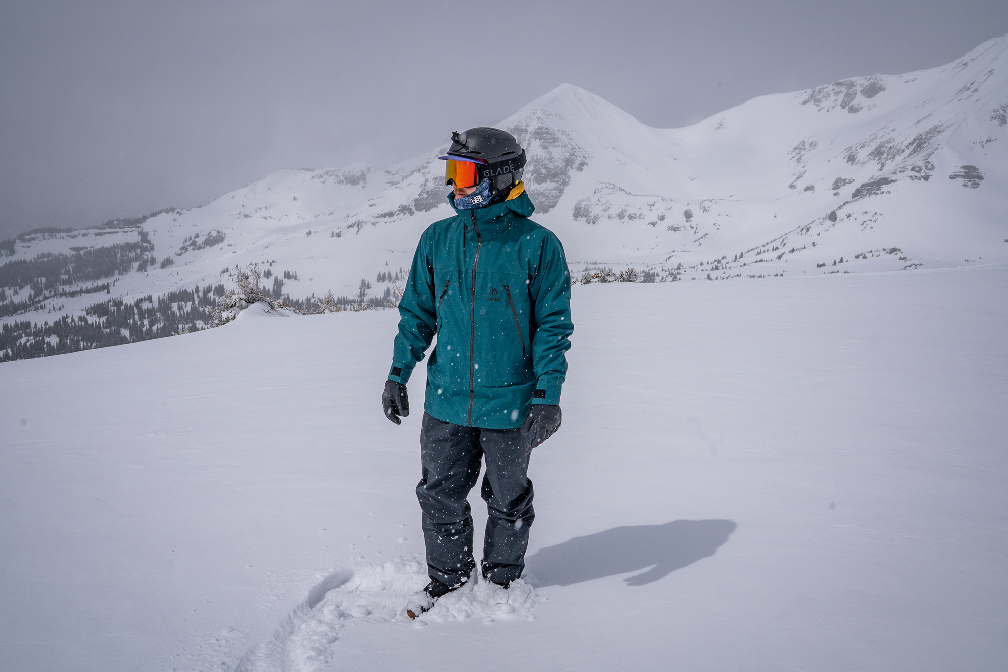 A snowboarder stands in fresh snow wearing the Jones Shralpinist jacket with mountains behind