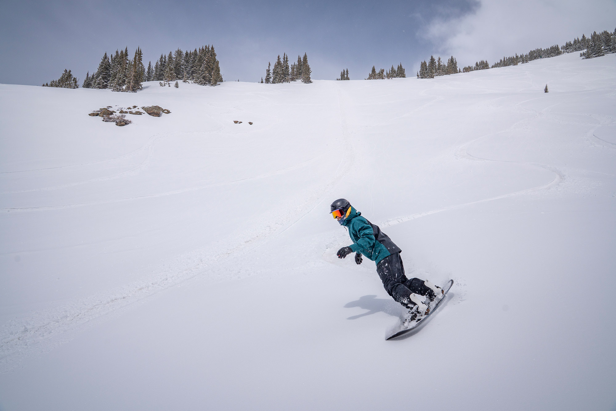 A snowboarder rides through soft powder wearing the Jones snowboard jacket