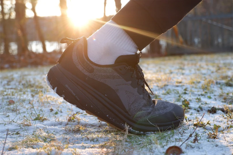 A dark athletic shoe lifting off the ground with snow and sunlight behind it