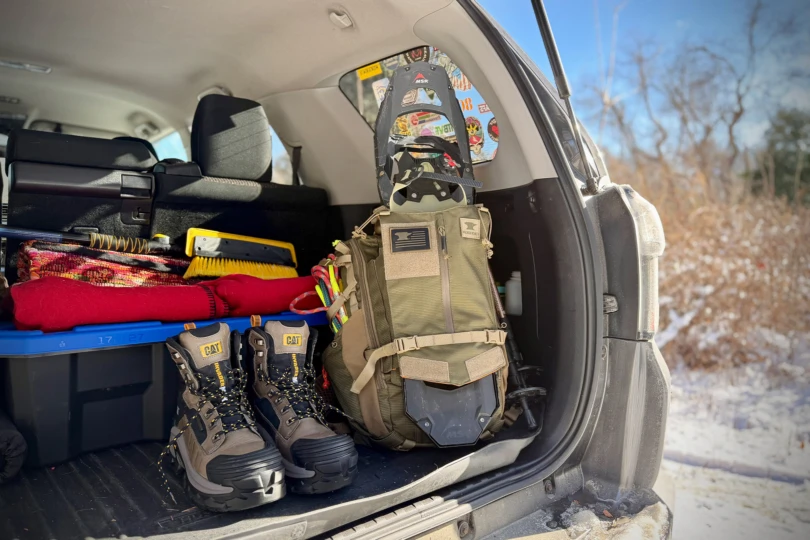 The backpack standing upright in a car trunk next to boots and winter gear