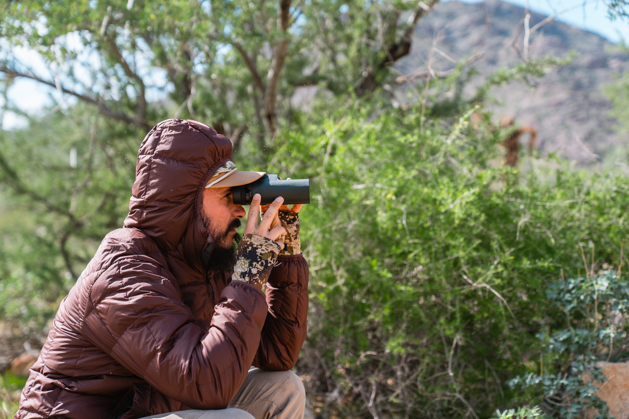A man in the Sitka down jacket kneels and looks through binoculars near thick desert shrubs