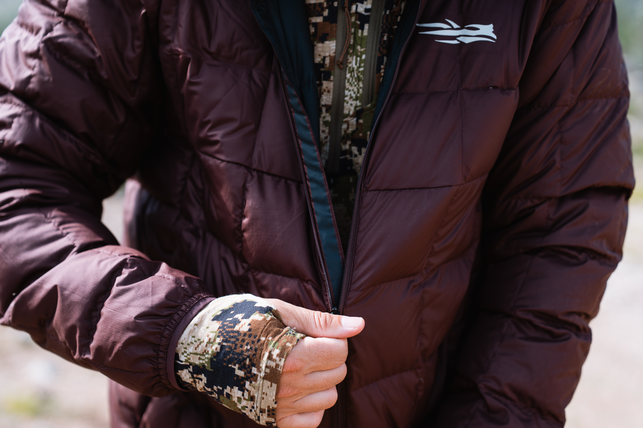 A close-up of someone zipping up the Sitka MicroDown Hooded Jacket, showing the quilted fabric and camo sleeve underneath