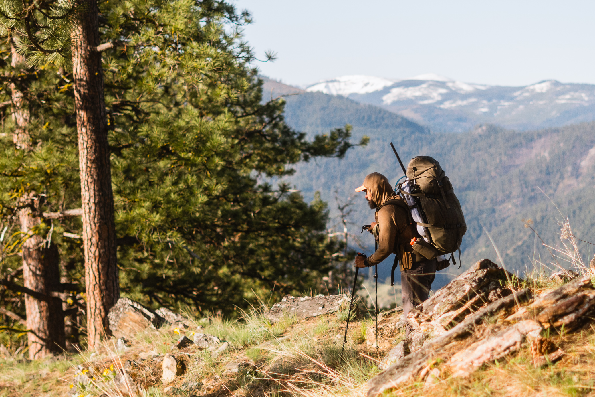 A man hikes uphill with trekking poles and a large pack while wearing the Sitka MicroDown Hooded Jacket in a forested mountain landscape