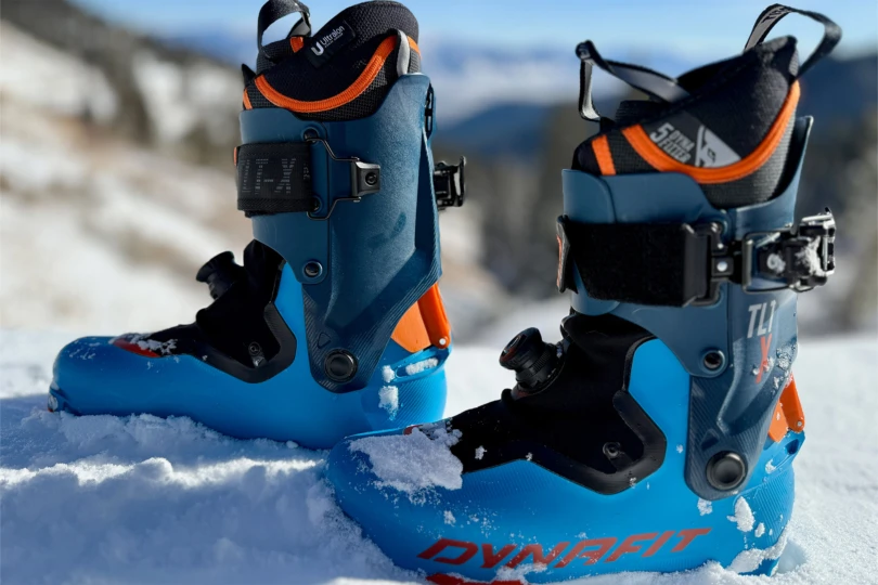 The touring boots stand in fresh snow with bright blue shells and orange details