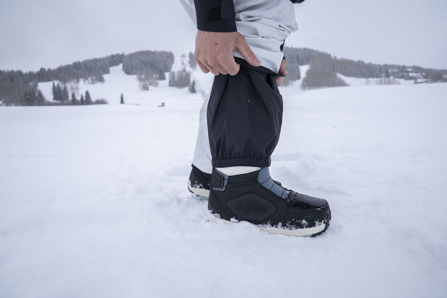A person standing in deep snow adjusts their pant gaiters over winter boots