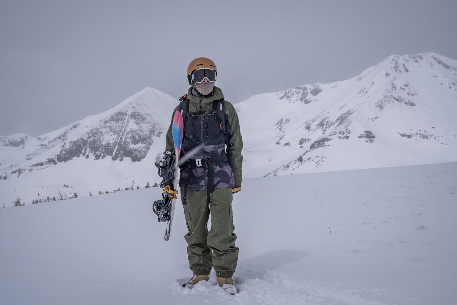 Snowboarder standing in deep snow with a board in hand, surrounded by snowy mountain peaks