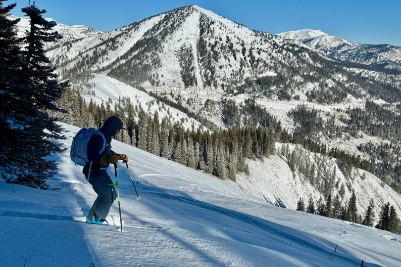 A skier pauses on a steep slope above a wide snowy valley