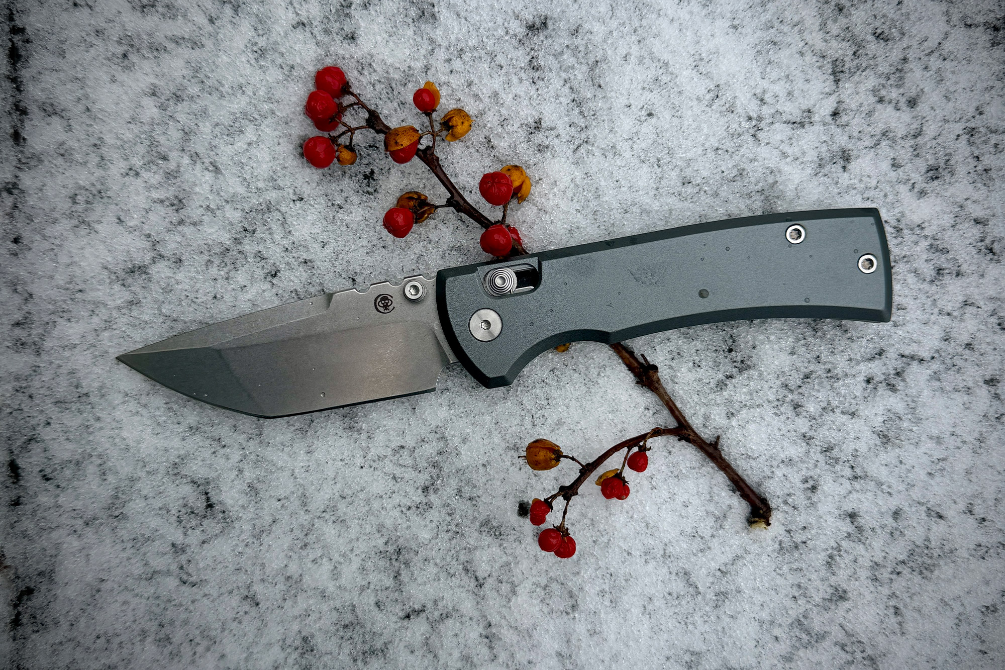 The knife laid open on snow with red berries nearby, highlighting the blade and handle finish