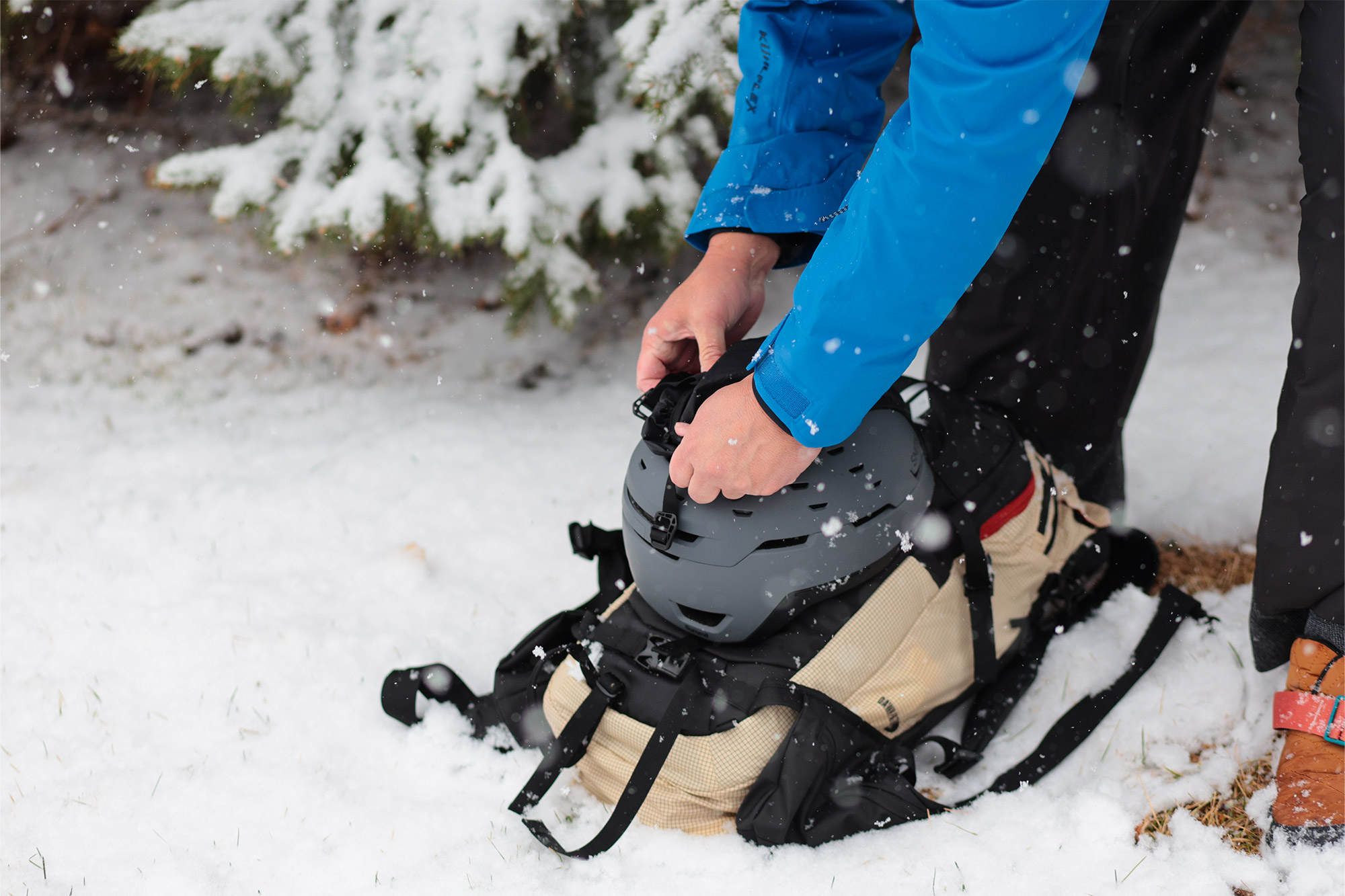 A helmet being attached to the Dawn Patrol ski pack while it rests on snow
