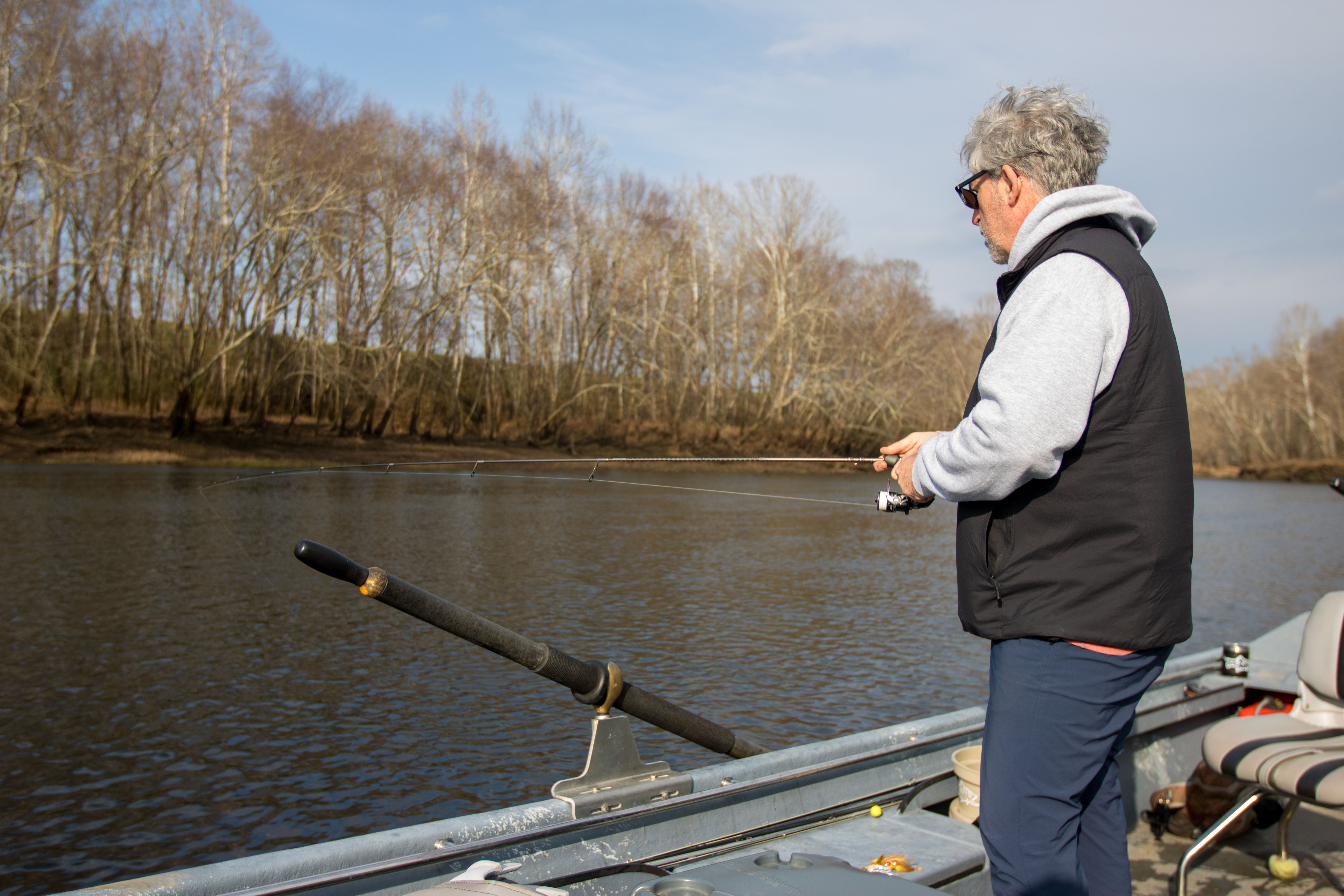 An angler fishing off the side of a small boat in a river.