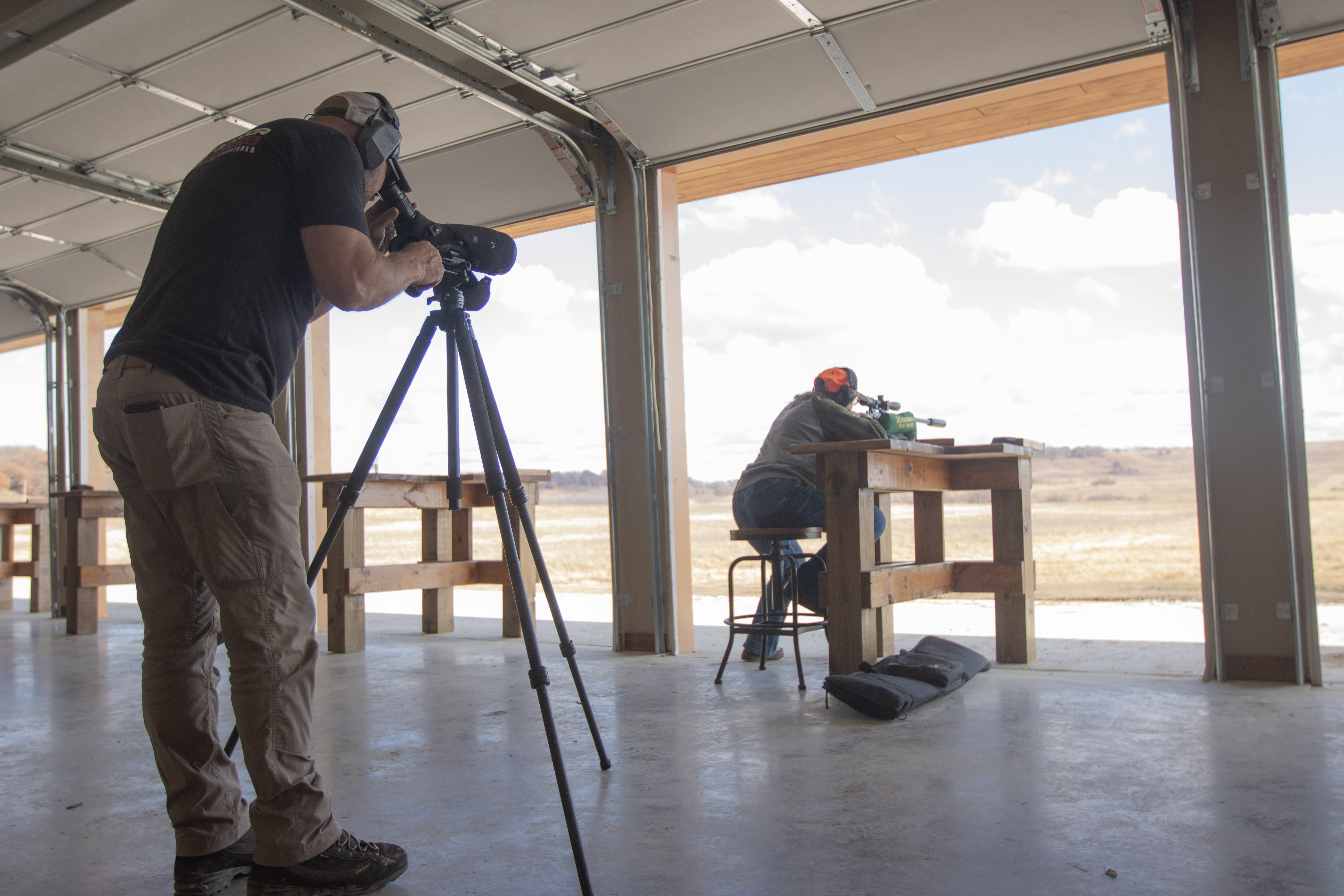 A woman shooting a rifle on a range while a spotter looks through a spotting scope to observe impacts.