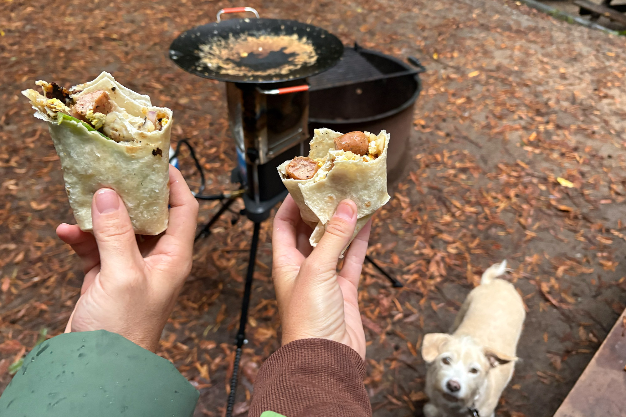 Two hands hold breakfast burritos in front of a campsite cooking setup, with the heater stove behind them and a small dog looking up from the ground