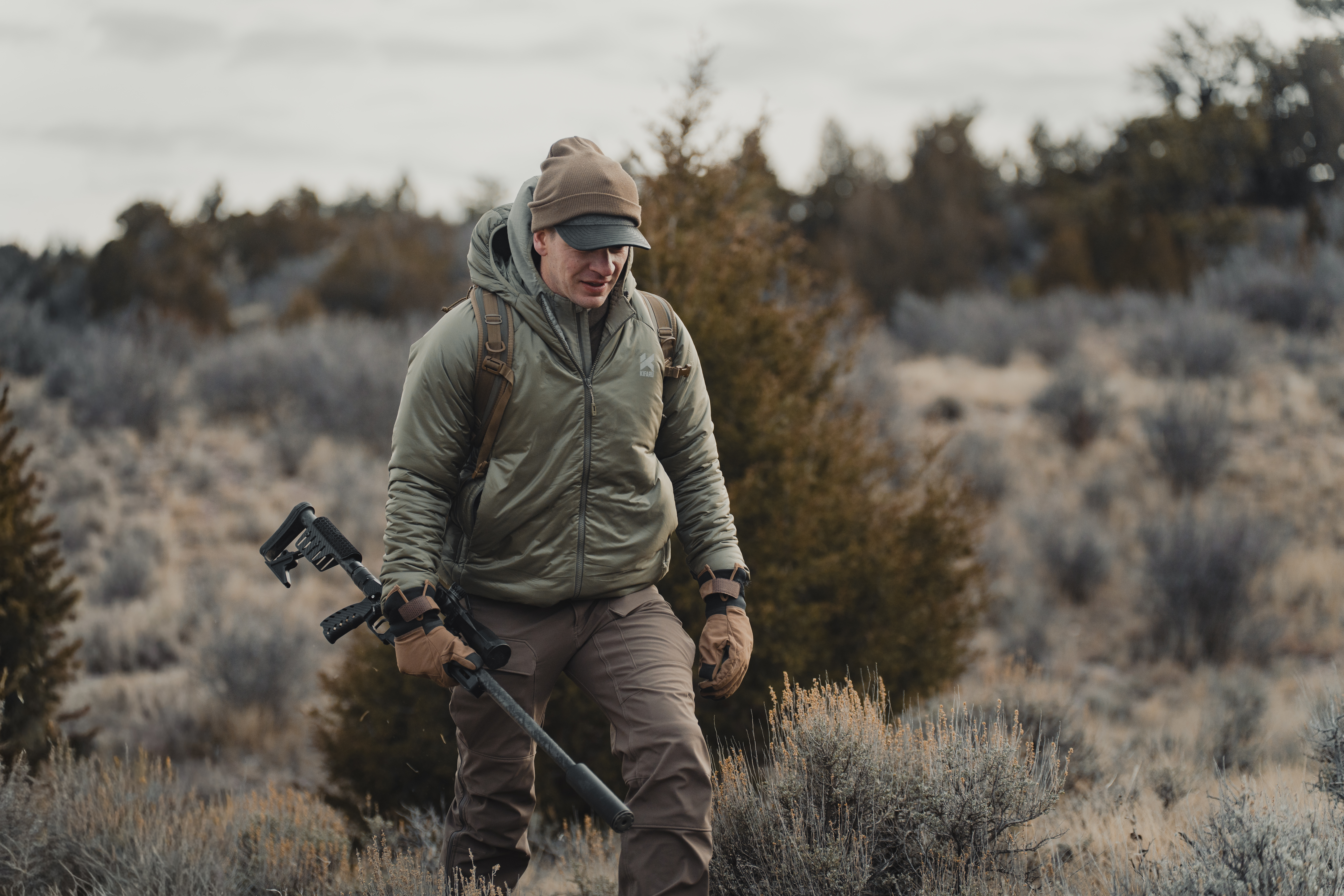 A hunter carrying a rifle walking through sagebrush.
