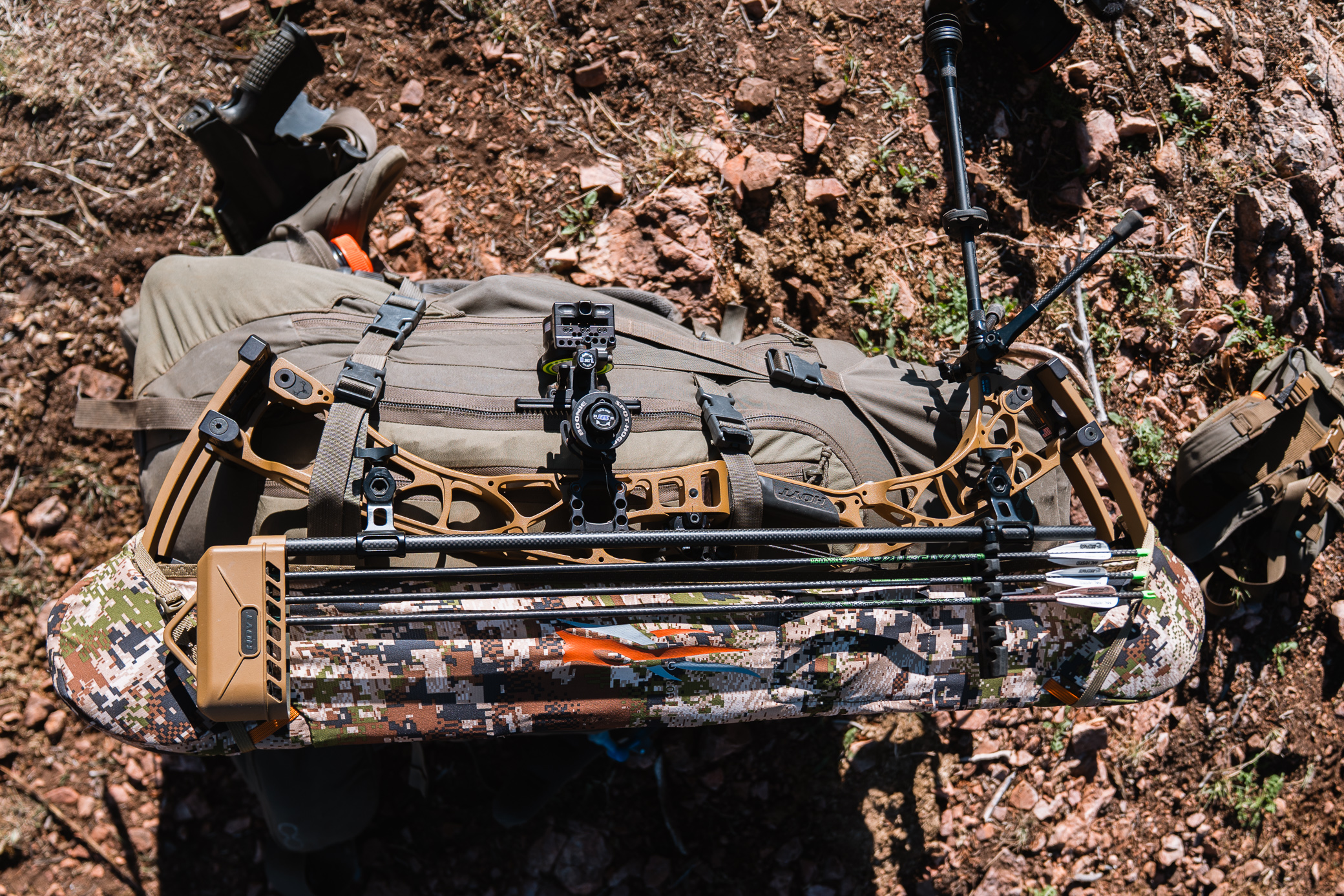A compound bow with the arrow rest secured is strapped to a hunting pack on rocky terrain