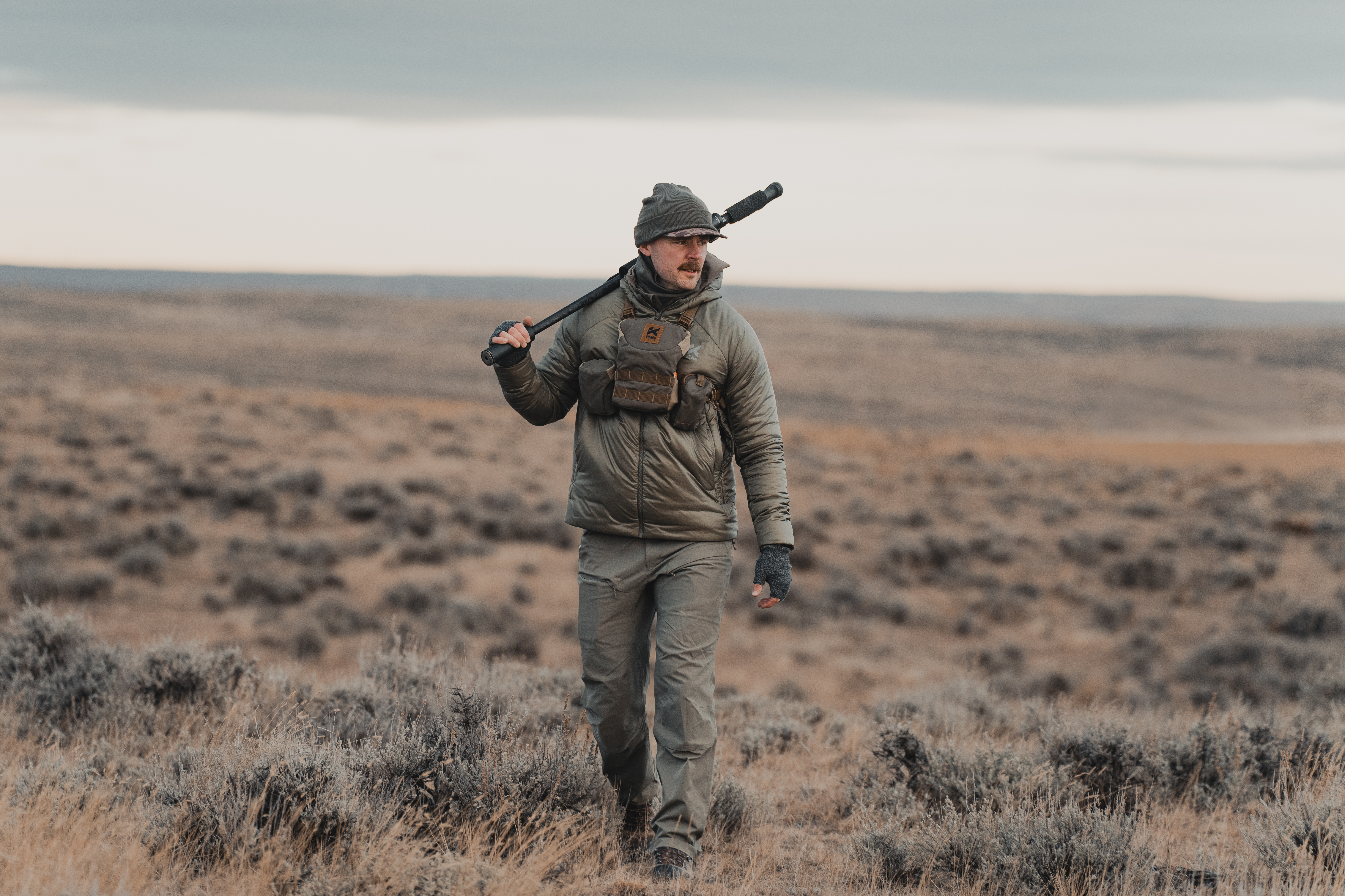 A hunter walking across a flat, sagebrush landscape wearing the new Kifaru apparel.