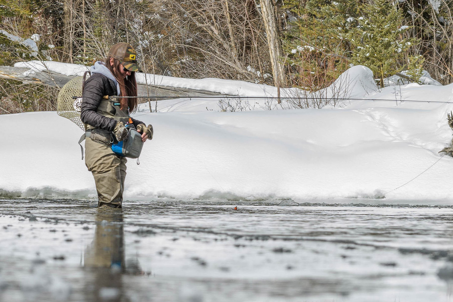 An angler stands in a shallow river surrounded by snow, wearing insulated PrimaLoft layers while preparing fishing gear