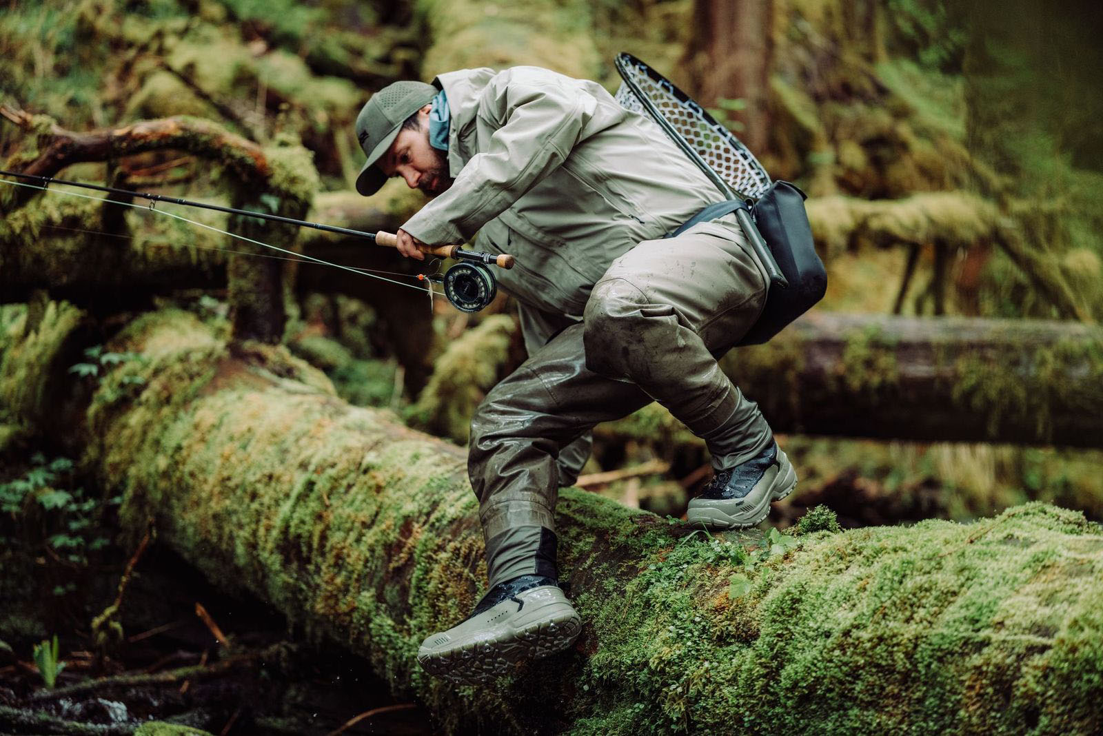 An angler in SITKA waders and boots climbing over a moss covered log while holding a fly rod.