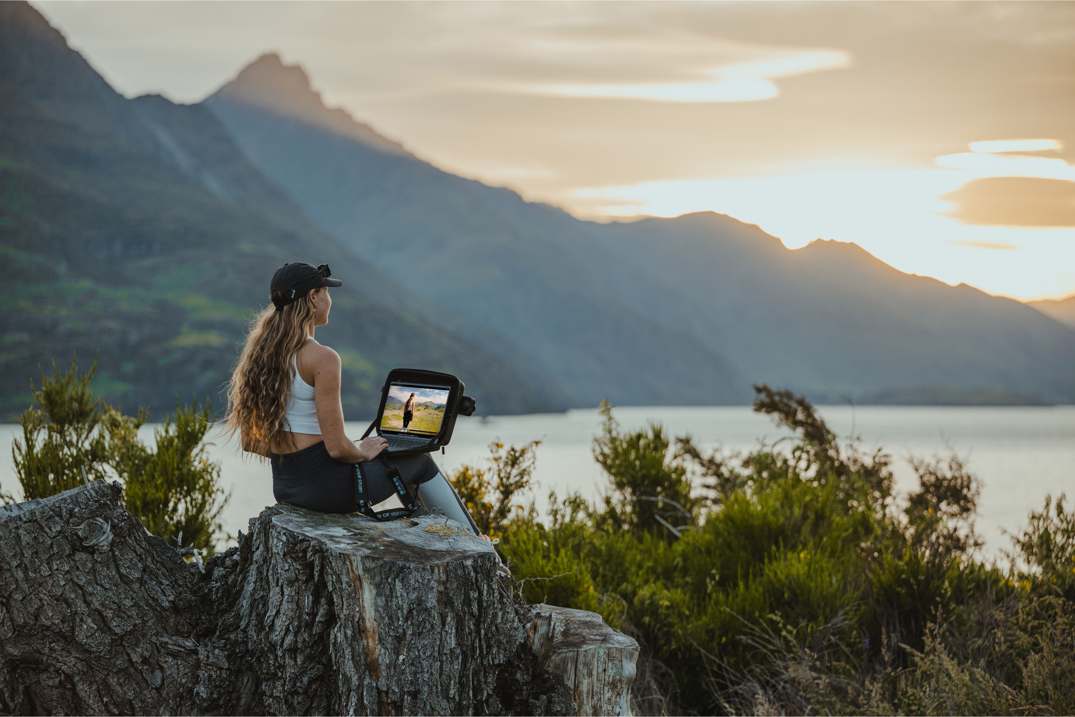 person sits by a lake and uses laptop