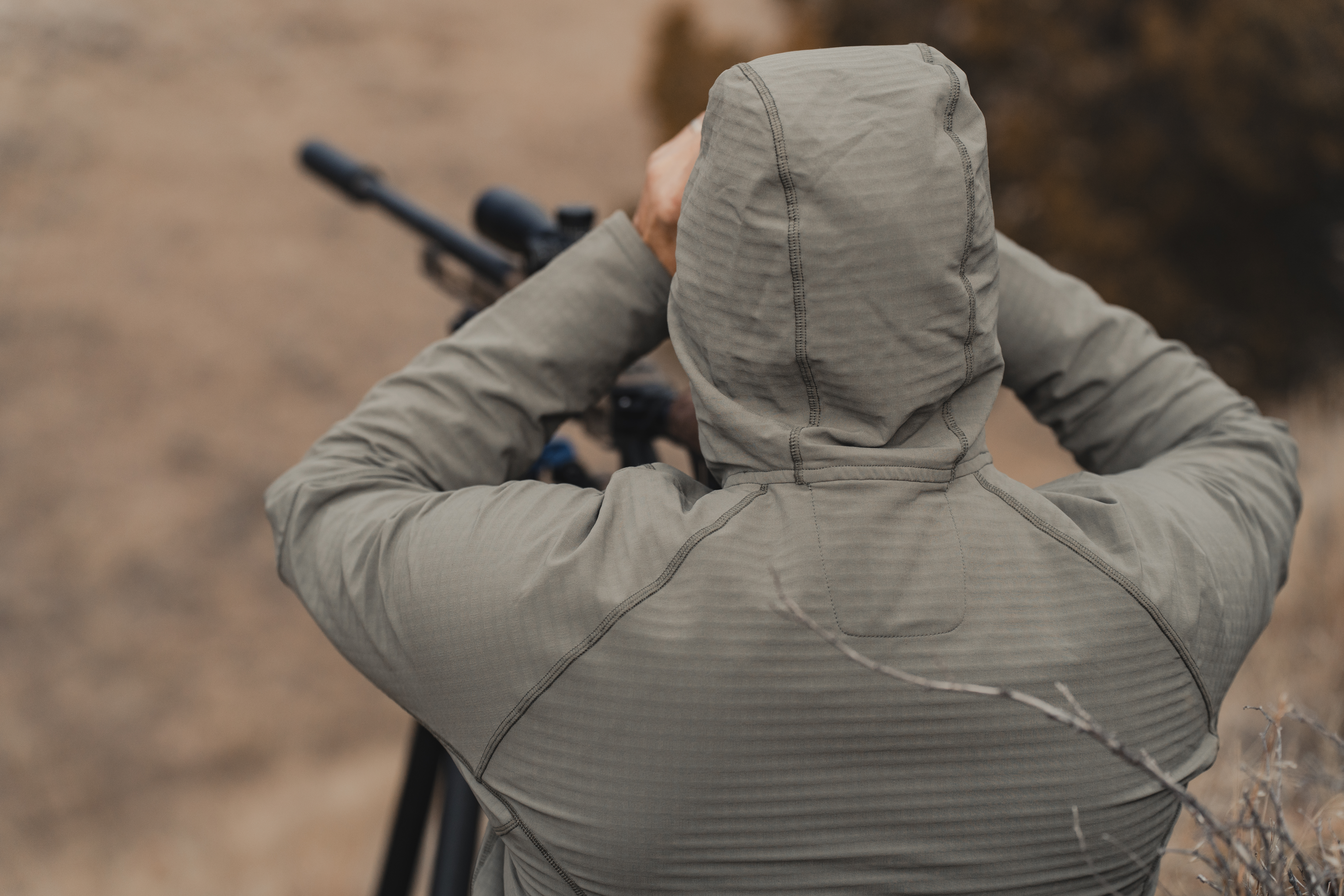The back of a hunter with a hood on as they glass behind a rifle and tripod