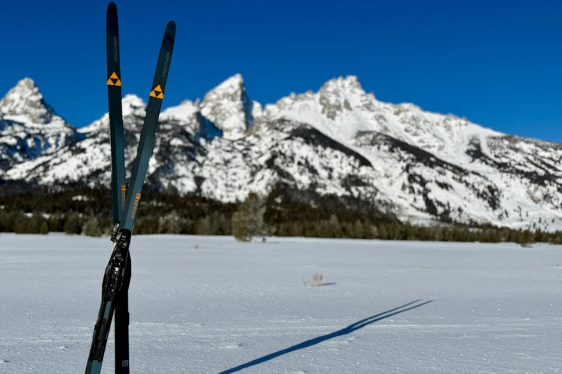 The Fischer Aeroguide 85 skis are planted upright in snow beneath clear mountain peaks
