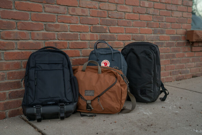 four of the best laptop backpacks sit on a sidewalk, including the timbuk2 authority, mystery ranch 3 way briefcase, fjallraven kanken, and evergoods cpl24