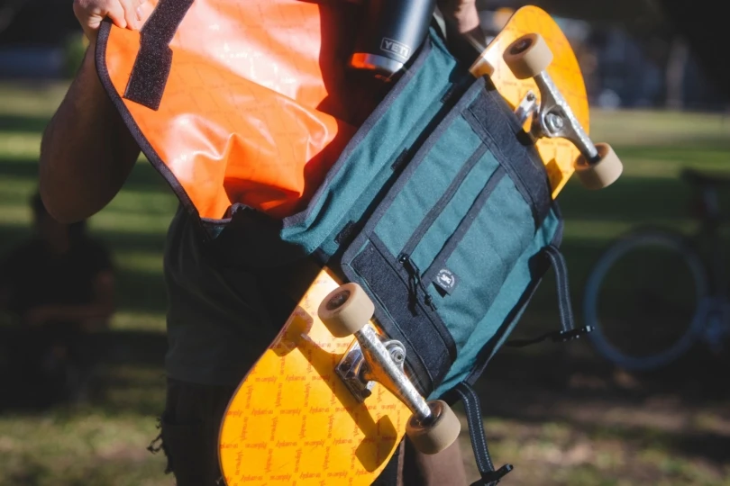 person puts water bottle in a messenger bag that has a skateboard on it