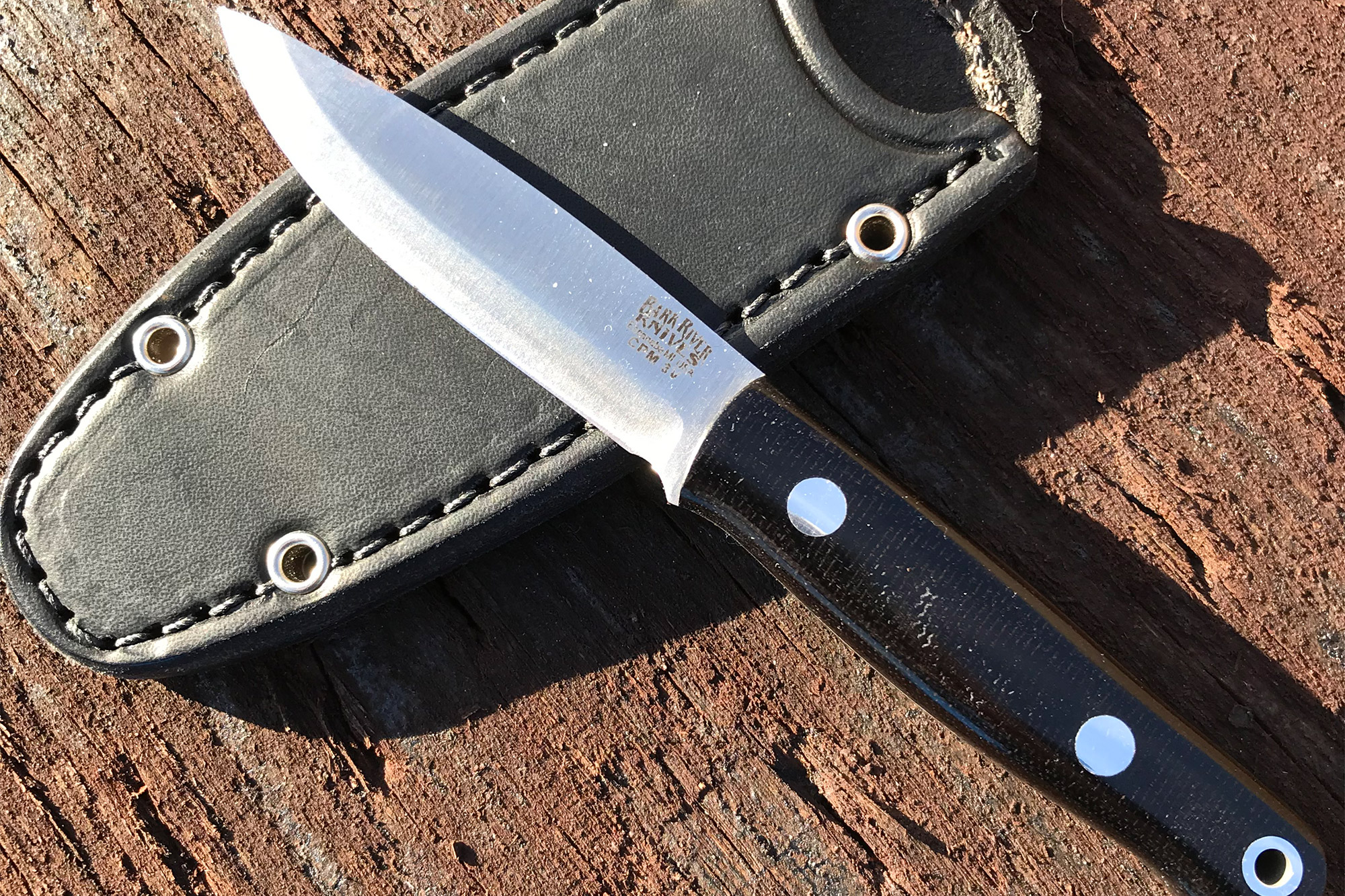 A small fixed blade knife rests on a black leather sheath on a wooden surface