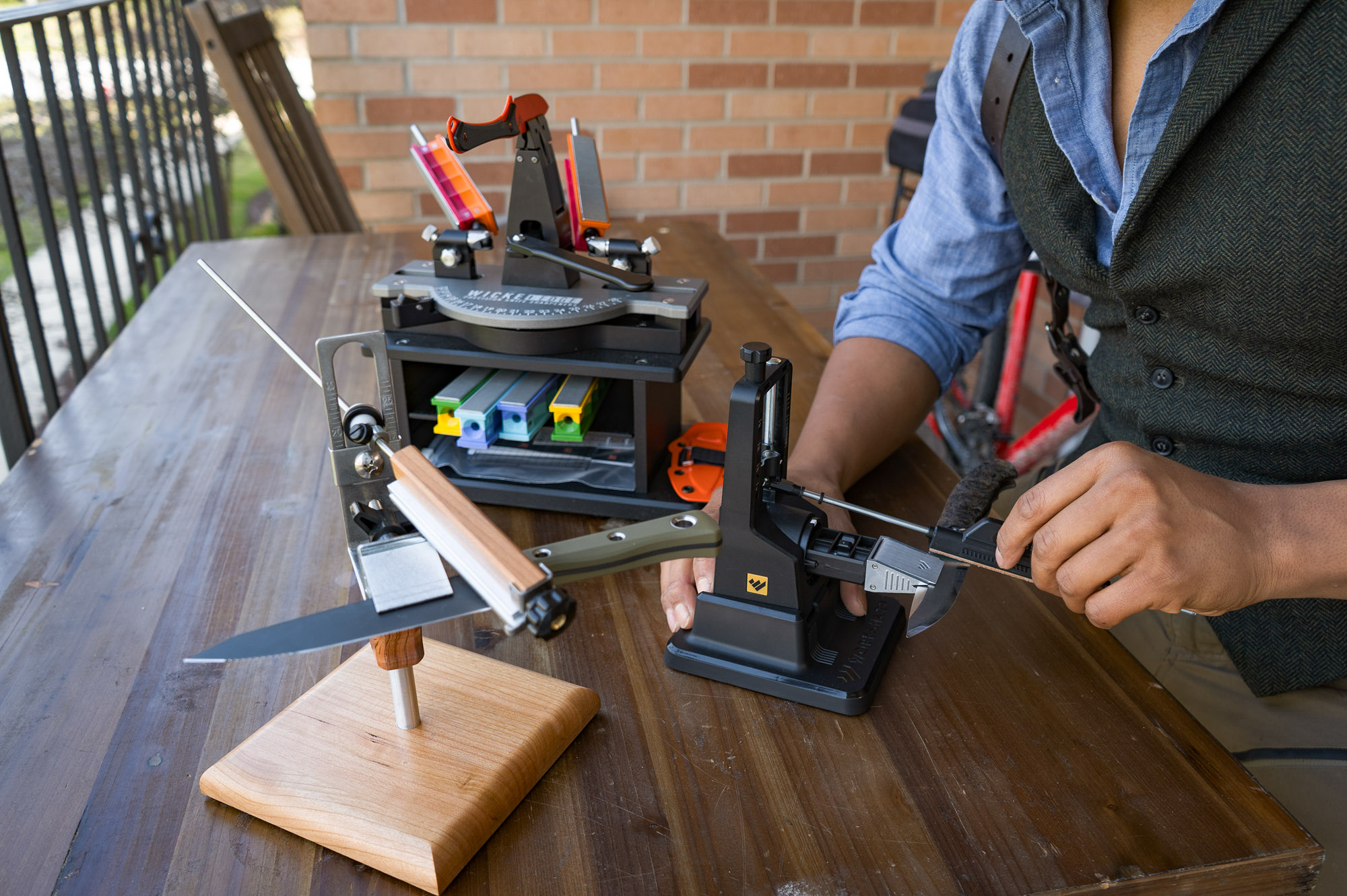 a trio of guided angle sharpeners on a benchtop being used to sharpen various pocket knives
